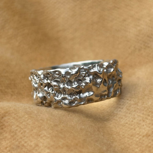 A textured sterling silver ring with an organic surface pattern, resting on a soft tan fabric background.